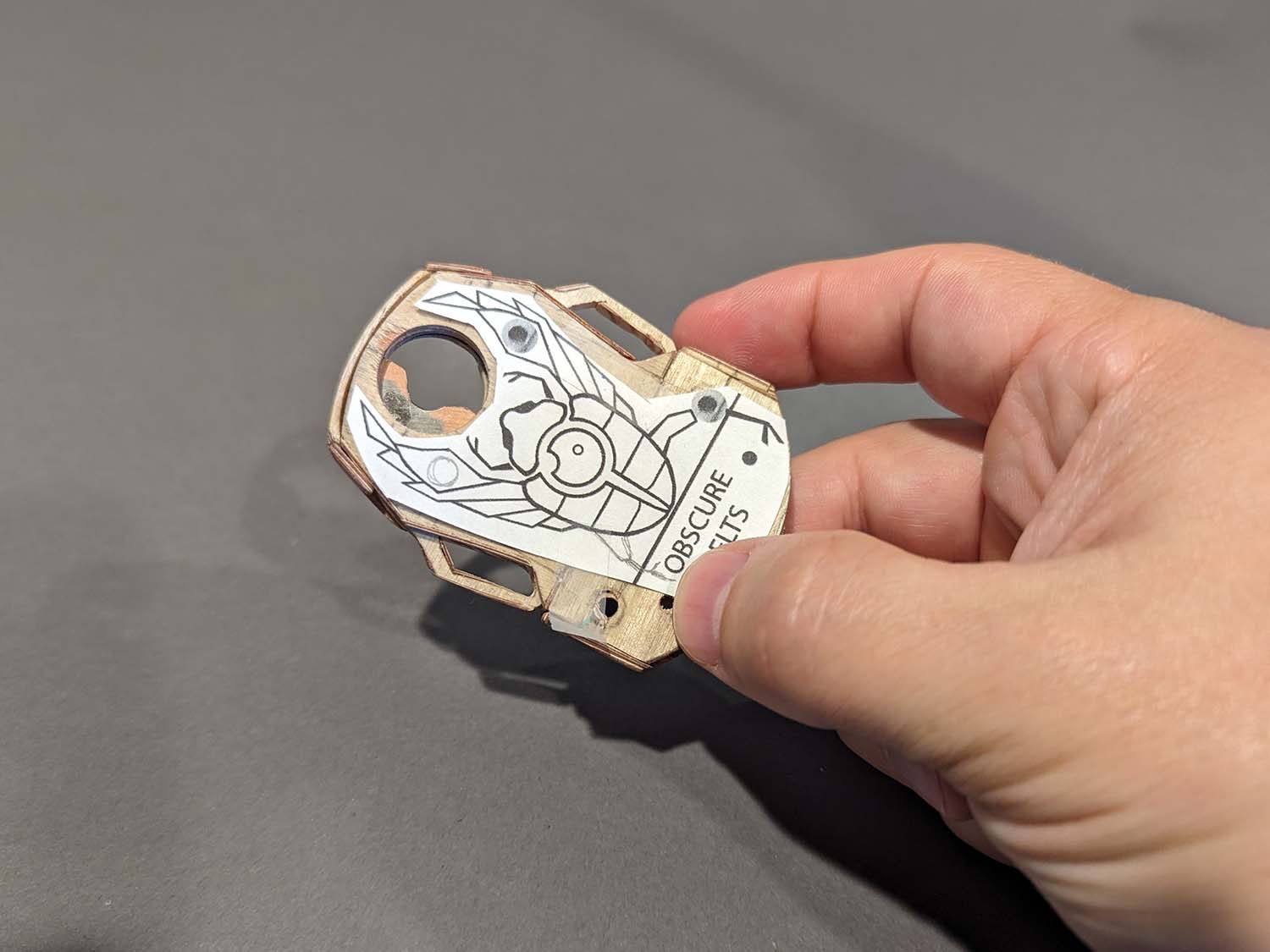 Hand holding a wooden prototype of mechanical belt buckle with scarab beetle shaped intricate design on a gray background