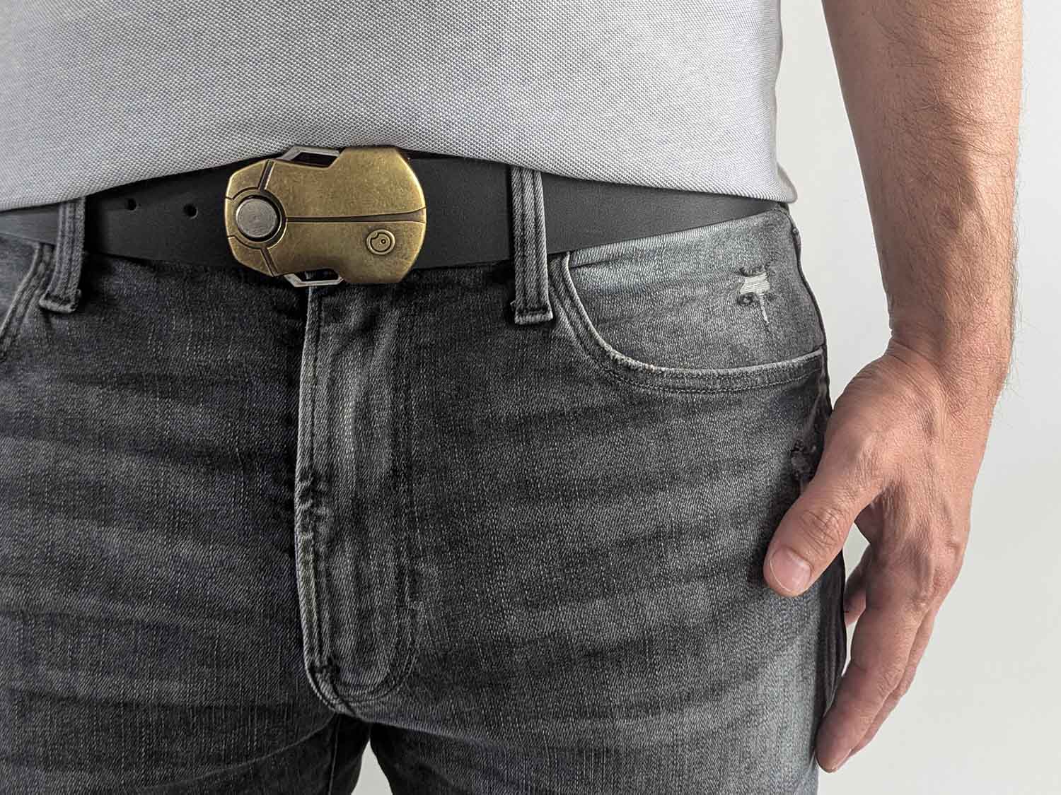 Person wearing a black leather belt with a gold buckle shaped like a scarab beetle on faded black jeans with a gray shirt against a white background