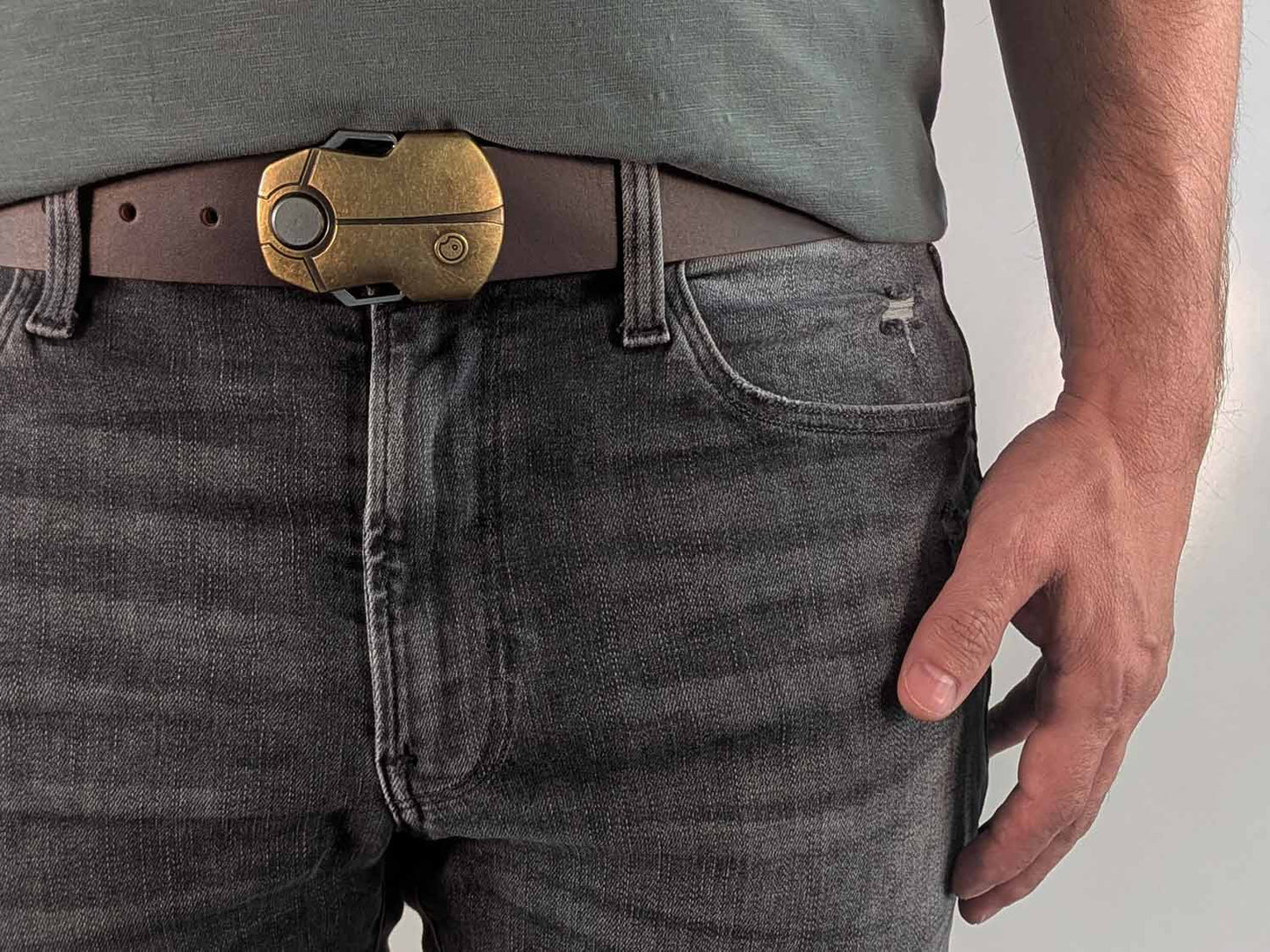 Person wearing a dark brown belt with a brass buckle shaped like a scarab beetle on faded black jeans with a green shirt against a white background