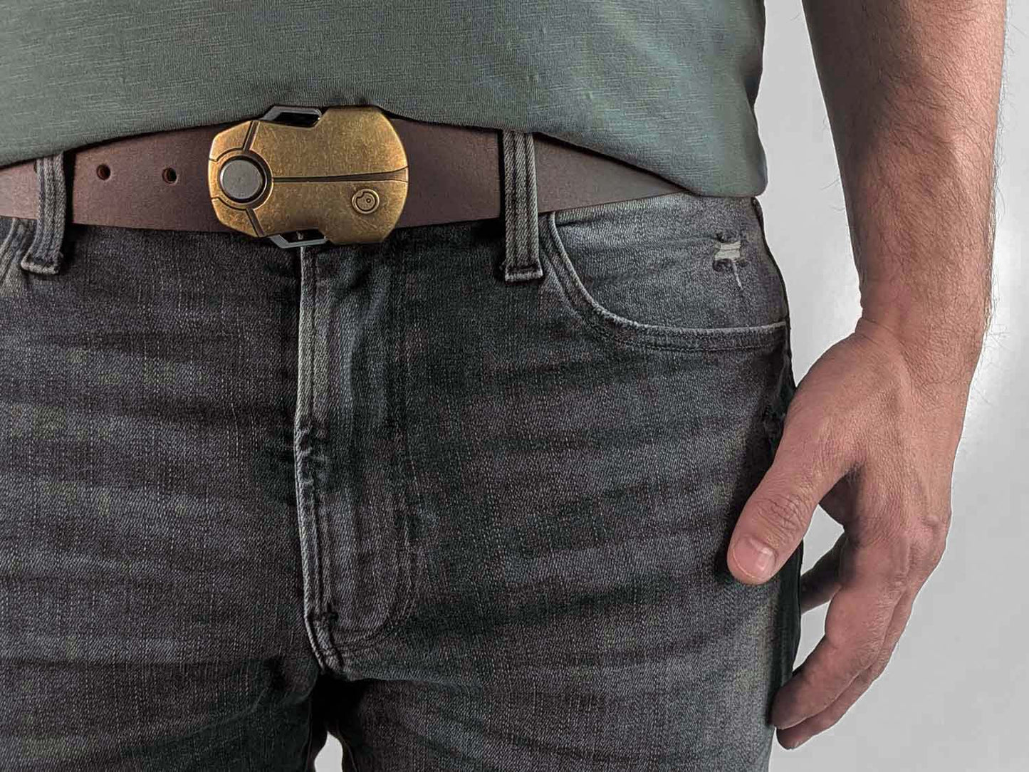 Person wearing a dark brown belt with a brass buckle shaped like a scarab beetle on faded black jeans with a green shirt against a white background