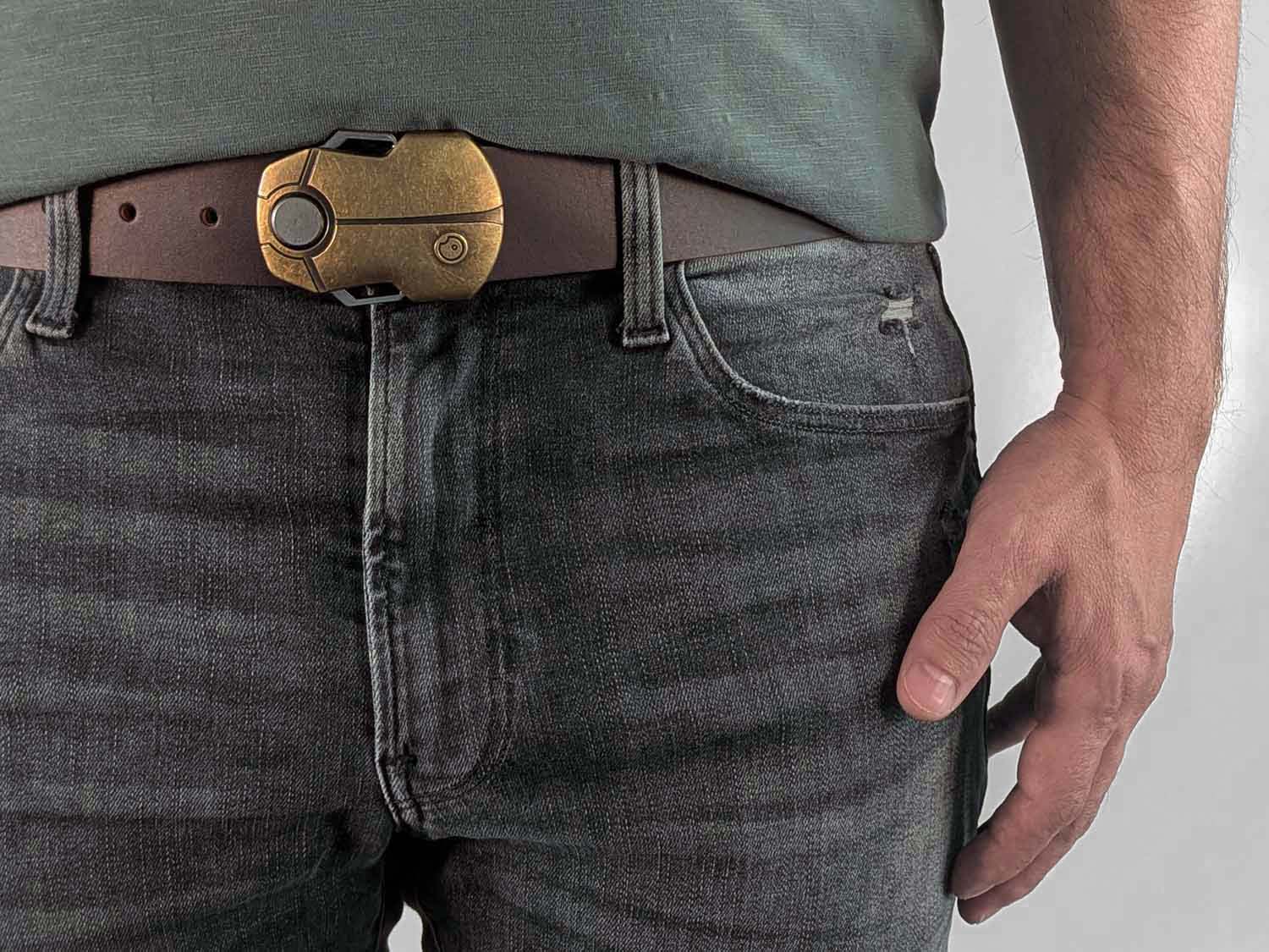 Person wearing a dark brown belt with a brass buckle shaped like a scarab beetle on faded black jeans with a green shirt against a white background