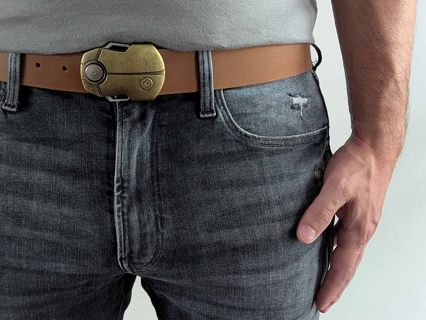 Person wearing a light brown leather belt with a black and gold buckle shaped like a scarab beetle on faded black jeans with a gray shirt against a white background