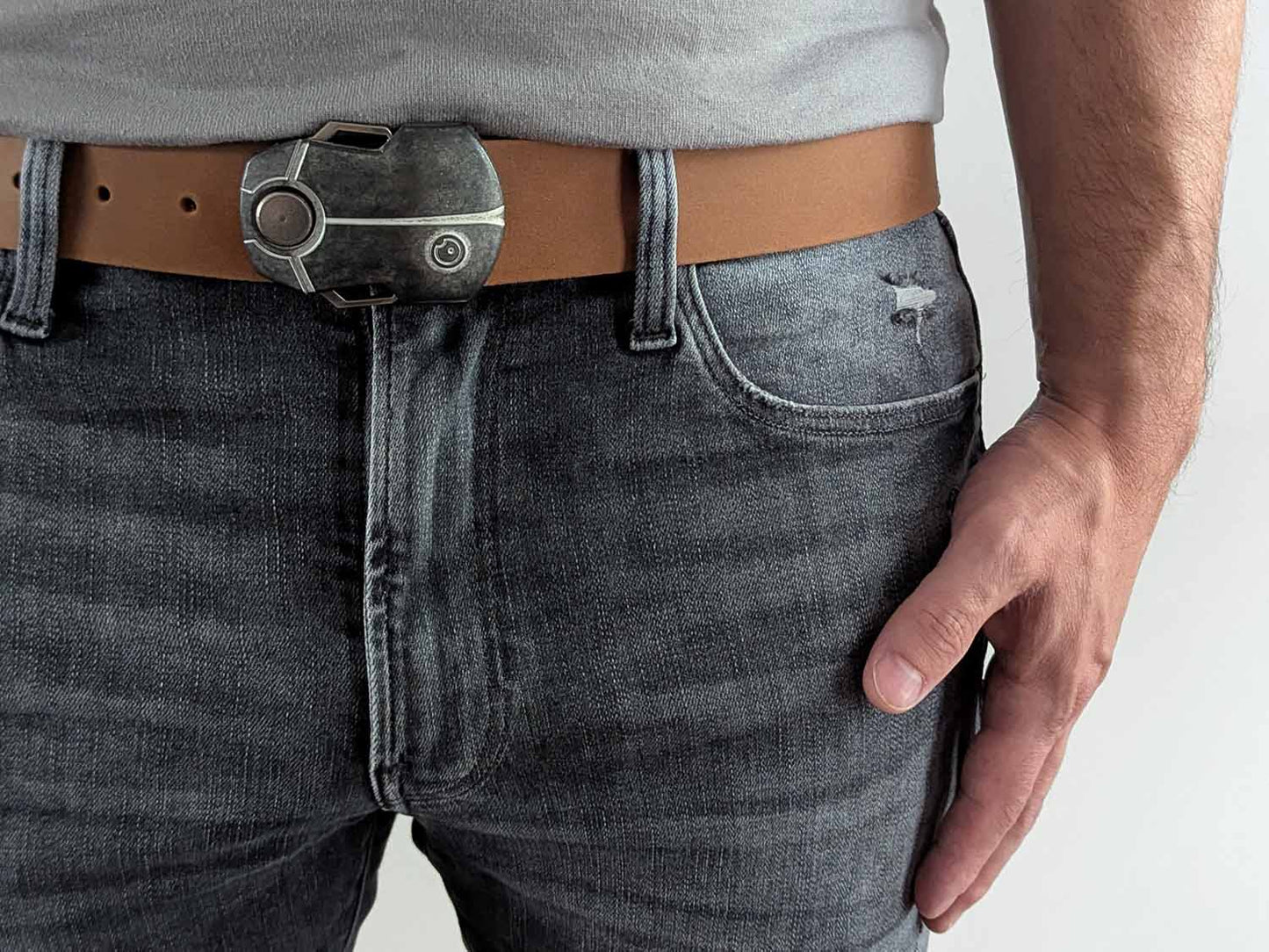 Person wearing a light brown belt with a metallic buckle shaped like a scarab beetle on faded black jeans with a gray shirt against a white background