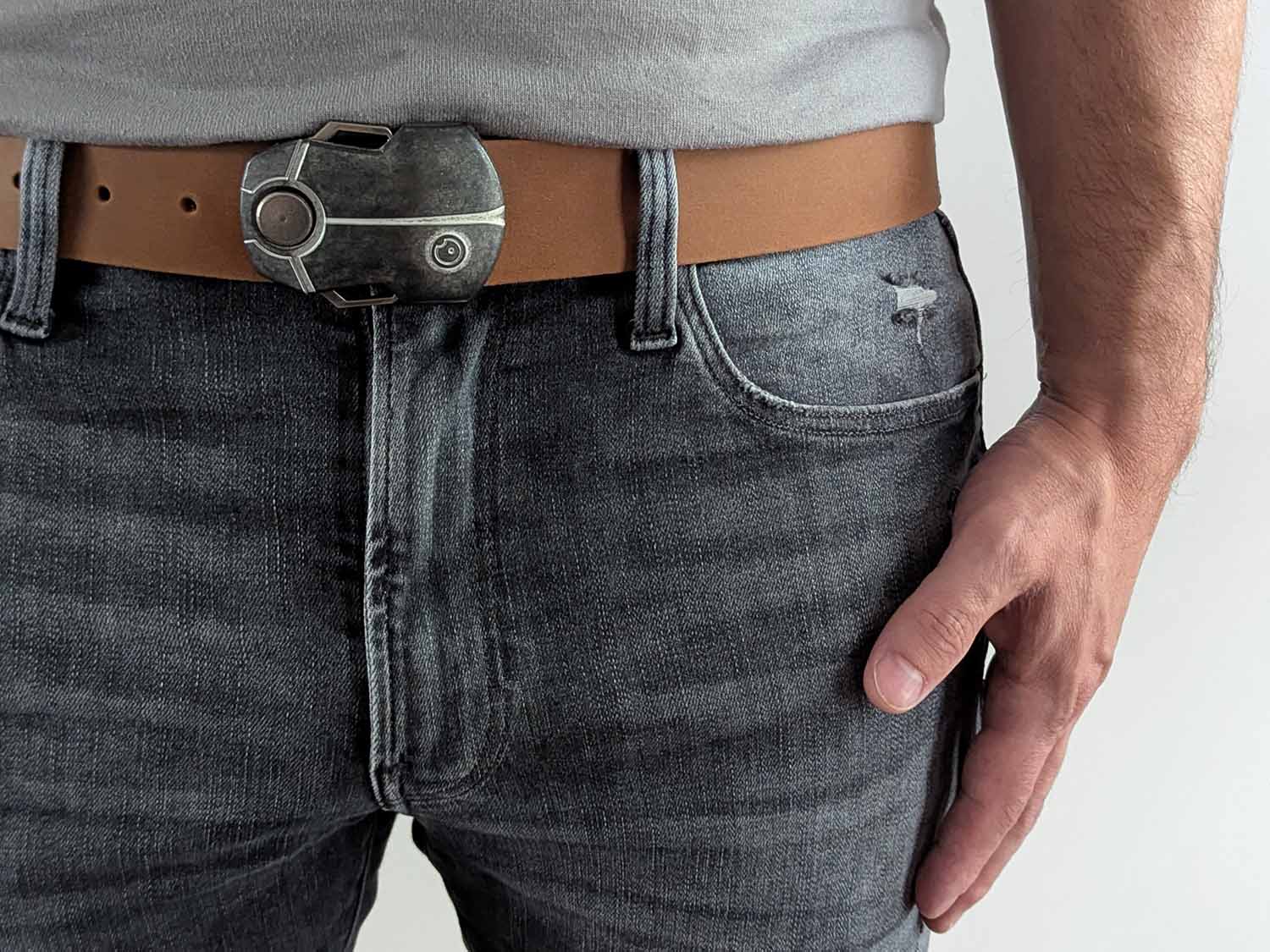 Person wearing a light brown belt with a metallic buckle shaped like a scarab beetle on faded black jeans with a gray shirt against a white background