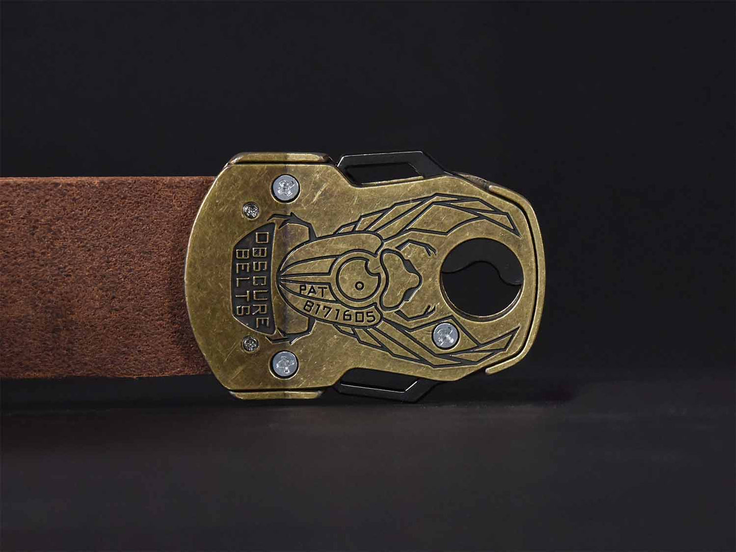 A magnetic belt buckle shaped like a scarab beetle with two satin silver wings and an antique brass buckle against a black background. The back of the gold belt buckle has a recessed scarab beetle design etched in black with the words "Obscure Belts" and the patent number.
