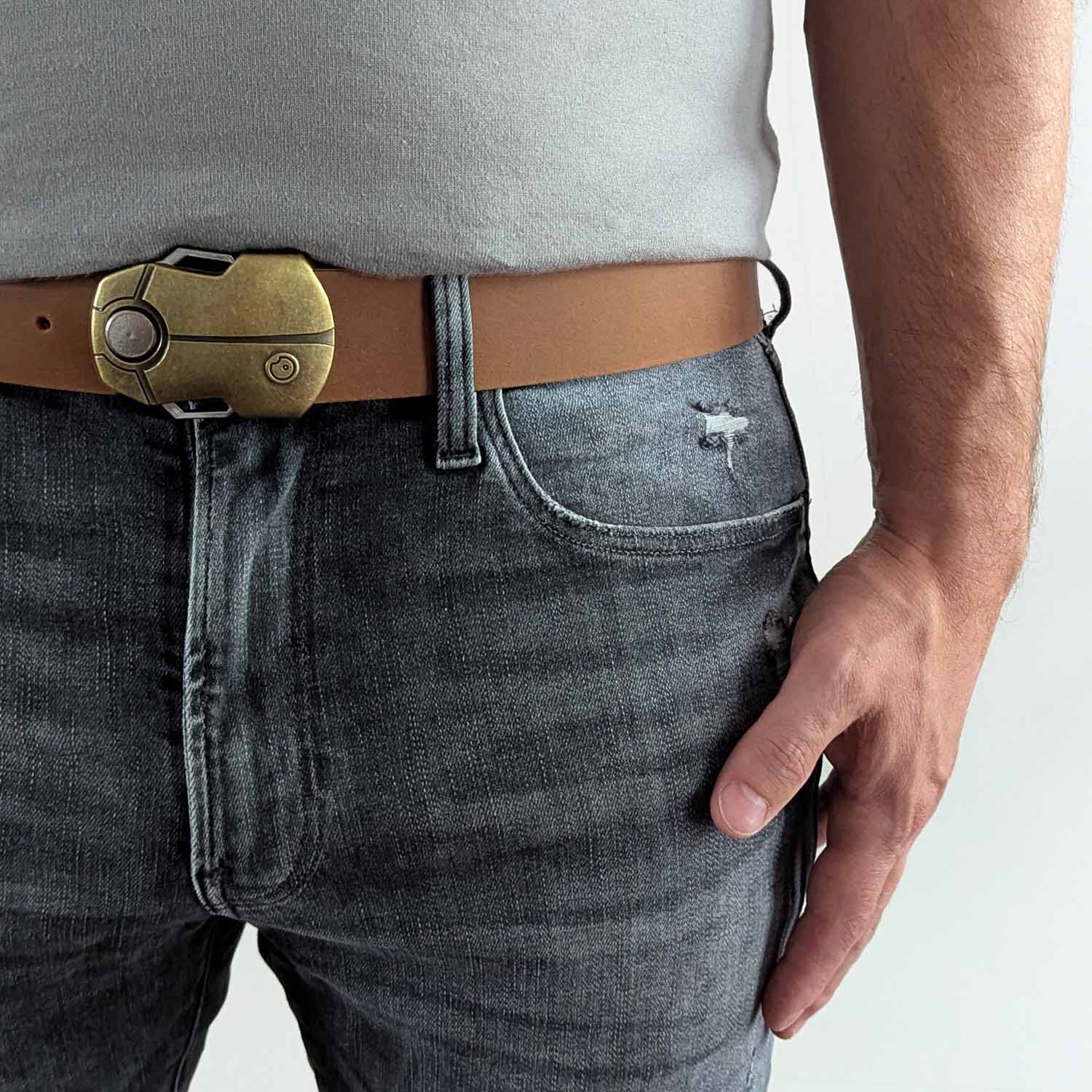 Person wearing a light brown leather belt with a black and gold buckle shaped like a scarab beetle on faded black jeans with a gray shirt against a white background