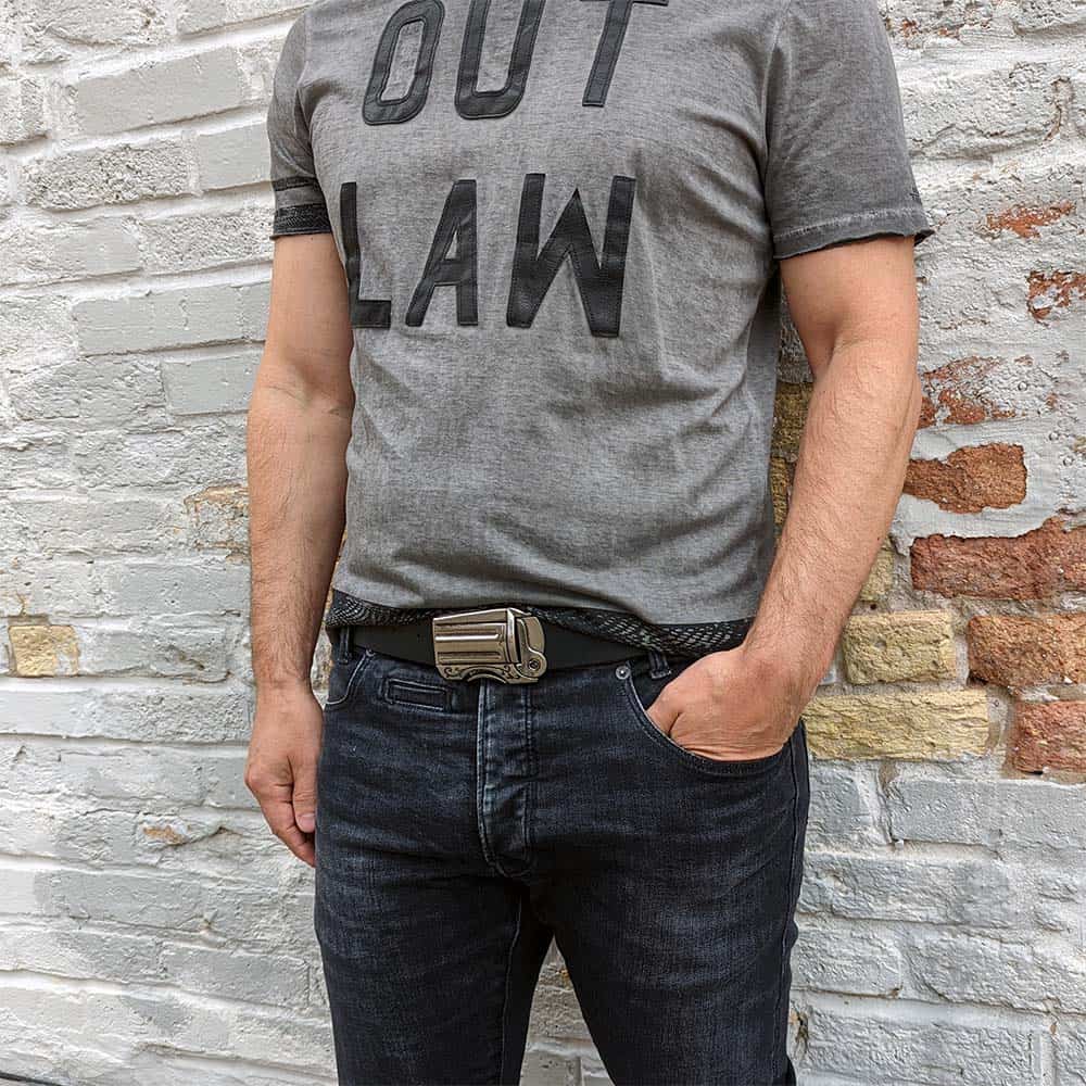 man wearing gun belt buckle with black jeans and a shirt that says OUTLAW against a grungy brick wall