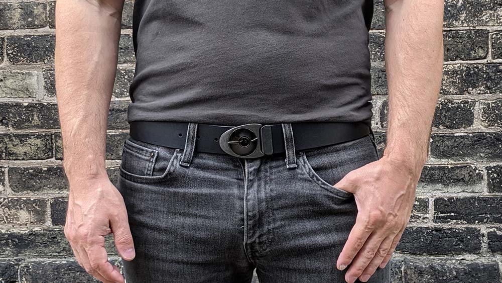 Person wearing a cool belt buckle on black leather belt with all black outfit again a dark brick wall. The belt has an industrial vibe.