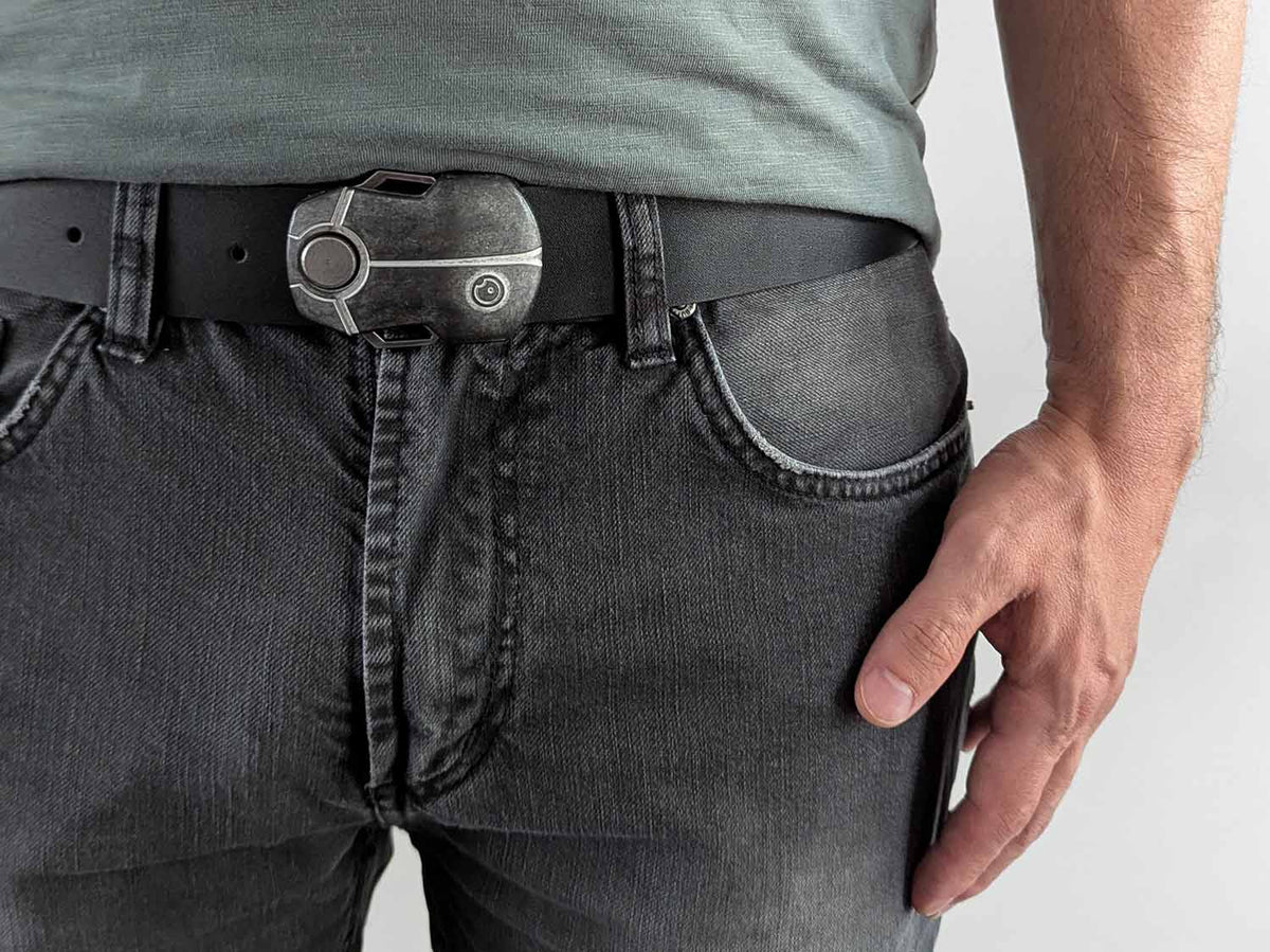 Person wearing a black belt with a metal buckle shaped like a scarab beetle on faded black jeans with a green shirt against a white background