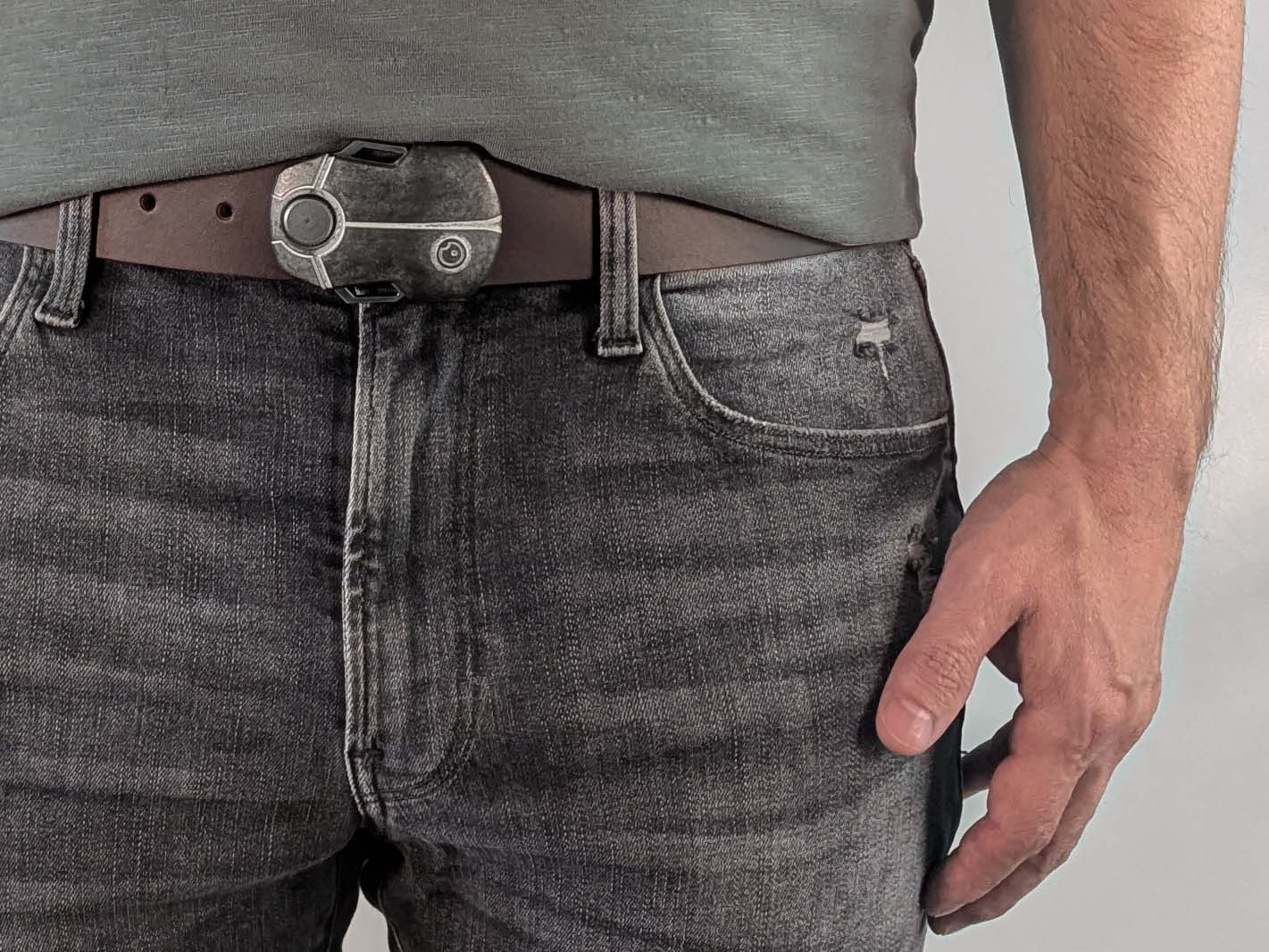 Person wearing a dark brown leather belt with a metallic buckle shaped like a scarab beetle on faded black jeans with a green shirt against a white background