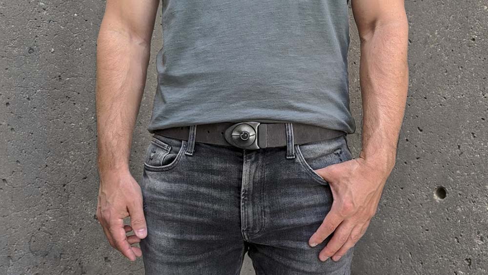 Person wearing unique arrowhead shaped belt buckle on a grey leather belt with a moss green shirt and faded grey jeans. Concrete wall in the background.