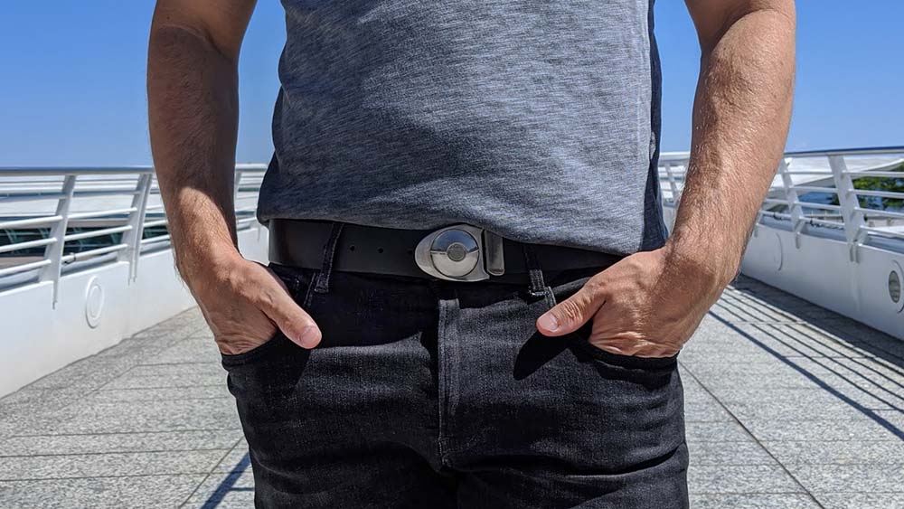 Person wearing unique silver arrowhead shaped belt buckle on a black leather belt with a heathered grey shirt and faded black jeans. Modern bridge and blue sky in the background.